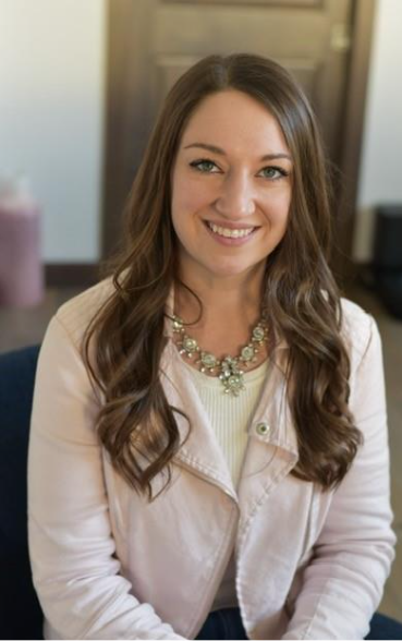 Casey sitting with brown hair pulled back and wearing glasses. Red professional top and gray bottom. Casey is smiling and looking at the camera.