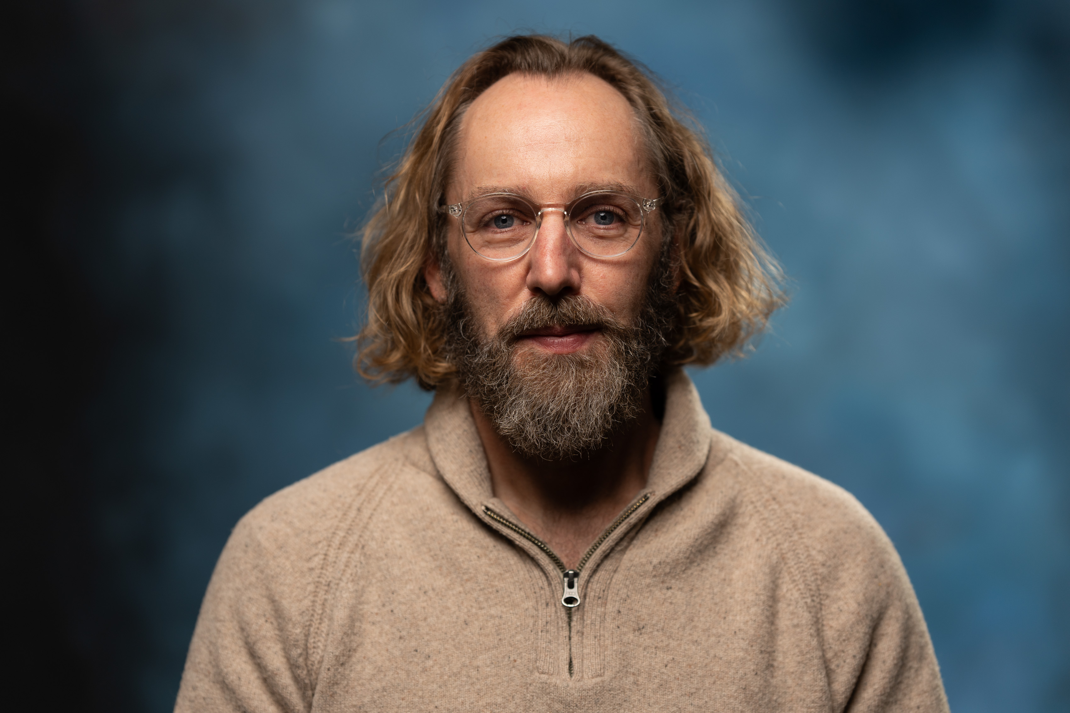 Joel Sands - Psychology Student of the Month, Joel is a white male wearing a brown sweater with a brown beard, brown hair, and glasses. There is a blue background.